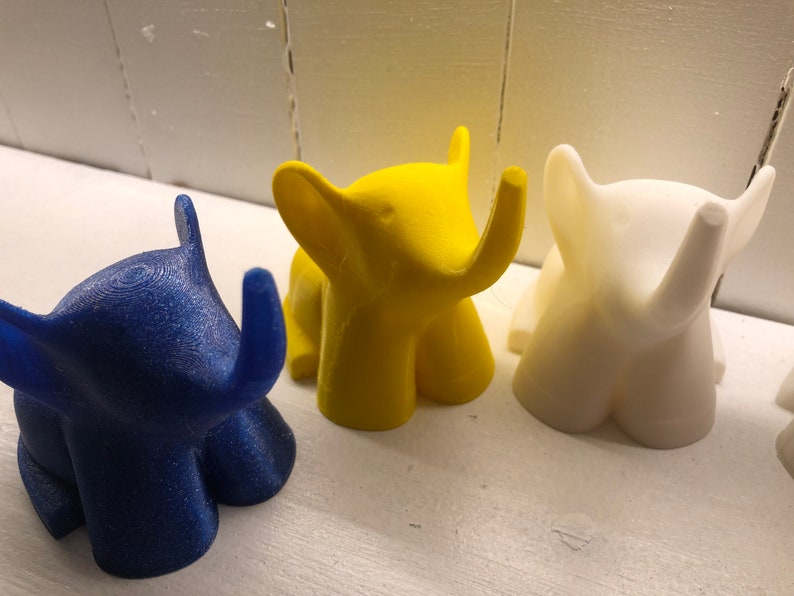 Op de afbeelding: Drie kleine olifantjes, een blauwe, een gele en een witte. De figuurtjes zijn gemaakt van plastic en hebben een gladde, glanzende afwerking.
