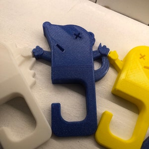 May include: Three 3D printed door handles in the shape of a cartoon character with a hook on the bottom. The handles are white, blue, and yellow.