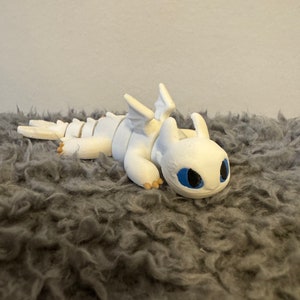 May include: White 3D printed figurine of a dragon with blue eyes. The dragon is lying on its belly with its wings spread out.