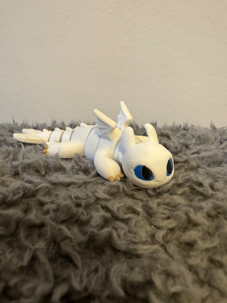 May include: White 3D printed figurine of a dragon with blue eyes. The dragon is lying on its belly with its wings spread out.