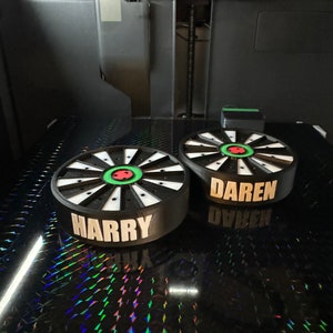 May include: Two custom dartboard-shaped objects with the names "HARRY" and "DAREN" printed in white. The dartboards have a black and white design with a green and red center. The objects are on a reflective surface, possibly being 3D printed.