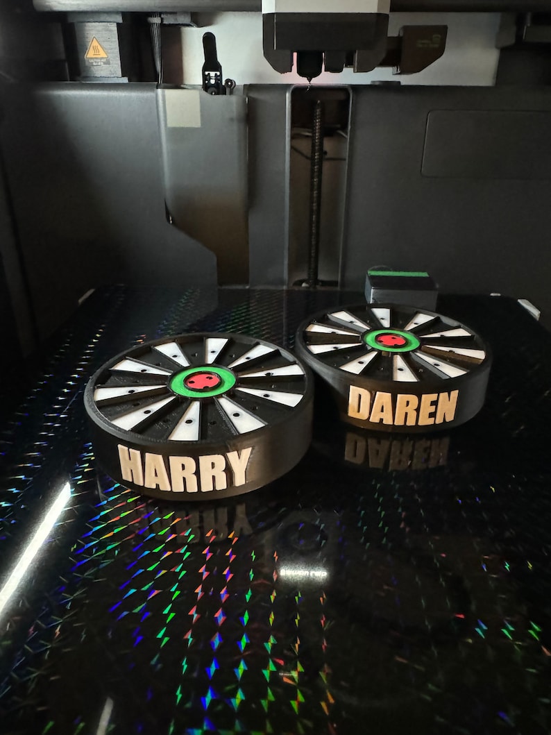 May include: Two custom dartboard-shaped objects with the names "HARRY" and "DAREN" printed in white. The dartboards have a black and white design with a green and red center. The objects are on a reflective surface, possibly being 3D printed.