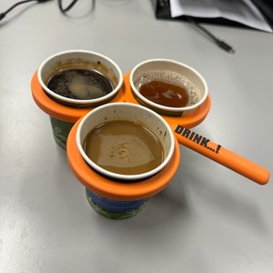 May include: Three cups of coffee in a portable orange holder with the text "DRINK...!".