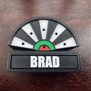 May include: A black and white dartboard with a green and red bullseye. The dartboard has a nameplate that reads "BRAD".