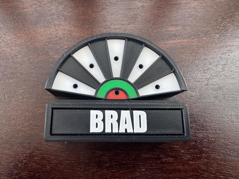 May include: A black and white dartboard with a green and red bullseye. The dartboard has a nameplate that reads "BRAD".