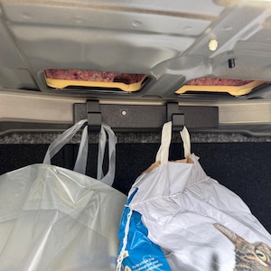 以下が含まれることがあります： 天井に取り付けられた黒い車のトランクカーゴフック。2つのビニール袋がフックから吊り下げられています。車の内装は明るいグレーで、下には濃いグレーのカーペットが敷かれています。