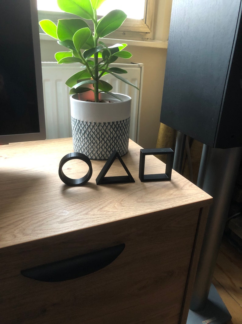 May include: Three black geometric shapes, a circle, a triangle, and a square, are arranged on a wooden surface. A potted plant with green leaves is in the background.
