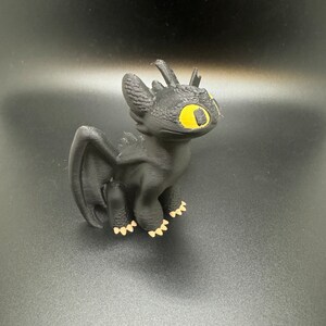 May include: Black 3D printed figurine of Toothless, a dragon from the How to Train Your Dragon franchise. The dragon has yellow eyes and is standing on a black surface.