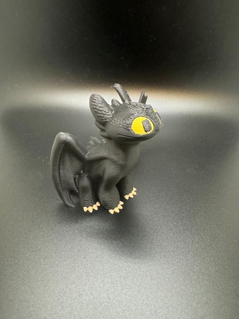 May include: Black 3D printed figurine of Toothless, a dragon from the How to Train Your Dragon franchise. The dragon has yellow eyes and is standing on a black surface.