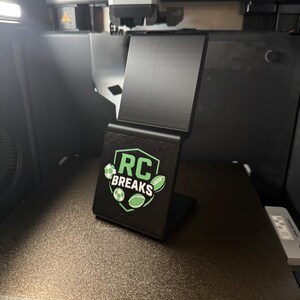 May include: A black 3D-printed phone stand with the text "RC BREAKS" and green sports ball graphics. The stand is angled and sits on a textured surface, likely inside a 3D printer.