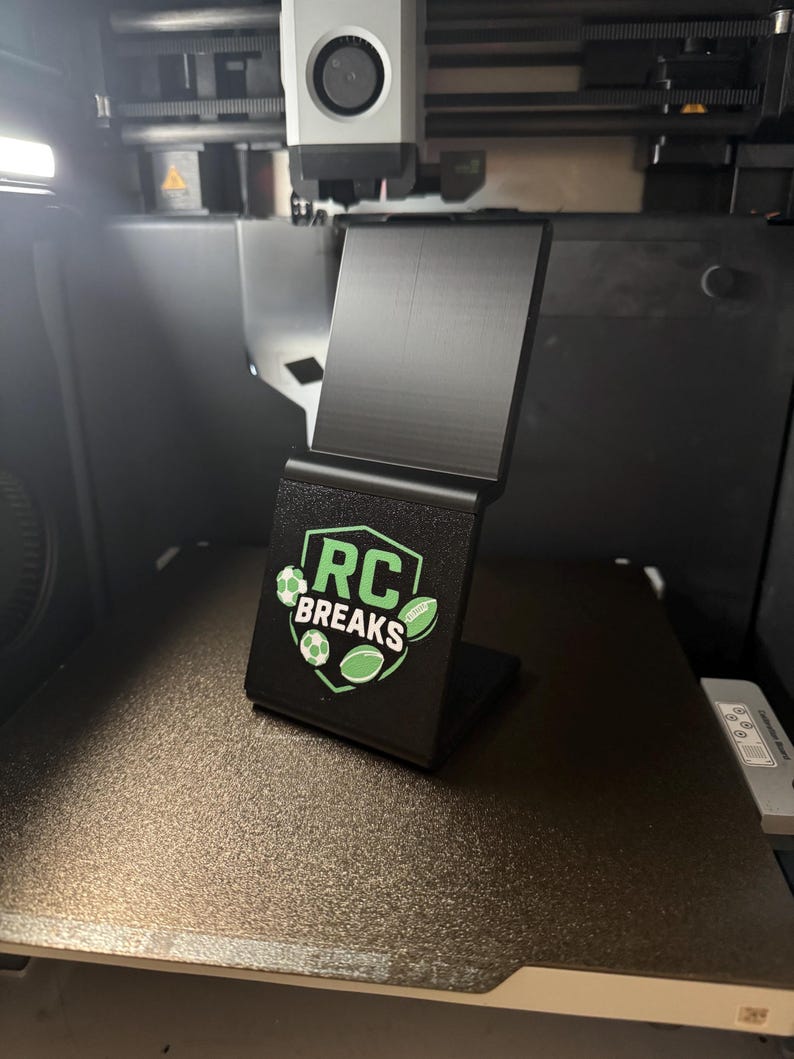 May include: A black 3D-printed phone stand with the text "RC BREAKS" and green sports ball graphics. The stand is angled and sits on a textured surface, likely inside a 3D printer.