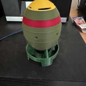 May include: A 3D-printed, multi-colored object resembling a bomb, featuring a yellow top, a red stripe, and an olive green body. It is set on a green base. The object has several small holes and is likely a decorative item or a model.