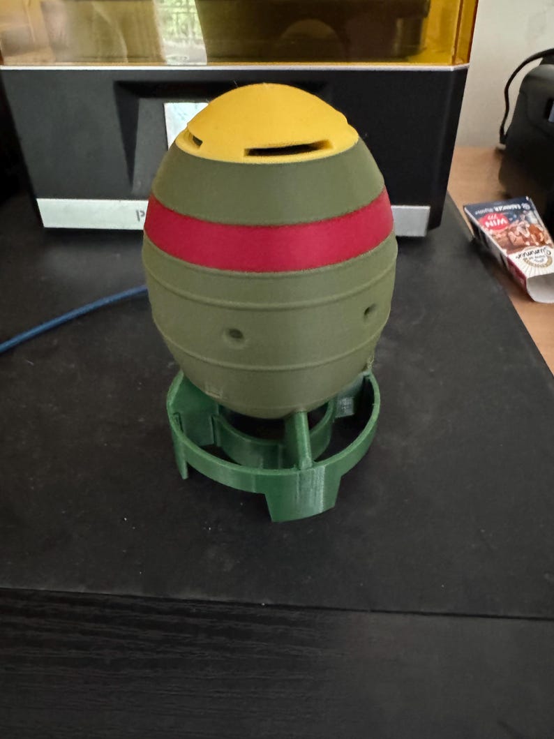 May include: A 3D-printed, multi-colored object resembling a bomb, featuring a yellow top, a red stripe, and an olive green body. It is set on a green base. The object has several small holes and is likely a decorative item or a model.