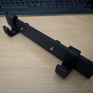 以下が含まれることがあります： 黒色の3Dプリントデバイス。長方形の本体と湾曲したフックがあり、電話やタブレットのホルダーの可能性があります。組み立て用のネジとスライドコンポーネントがあります。デバイスは明るい木目調の表面にあります。