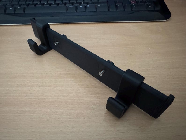 以下が含まれることがあります： 黒色の3Dプリントデバイス。長方形の本体と湾曲したフックがあり、電話やタブレットのホルダーの可能性があります。組み立て用のネジとスライドコンポーネントがあります。デバイスは明るい木目調の表面にあります。