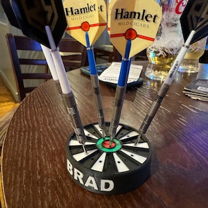 May include: A set of darts in a black and white holder with the name "BRAD" printed on the side. The darts have various colored flights, including black, yellow, and blue. The darts are arranged on a wooden table.