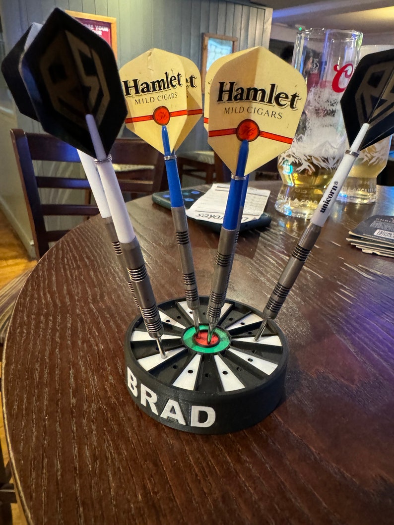 May include: A set of darts in a black and white holder with the name "BRAD" printed on the side. The darts have various colored flights, including black, yellow, and blue. The darts are arranged on a wooden table.