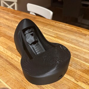 May include: A black, 3D-printed Darth Vader helmet-shaped phone holder. The holder has a textured surface and a slot to hold a phone. The design is detailed and the holder is sitting on a wooden table.