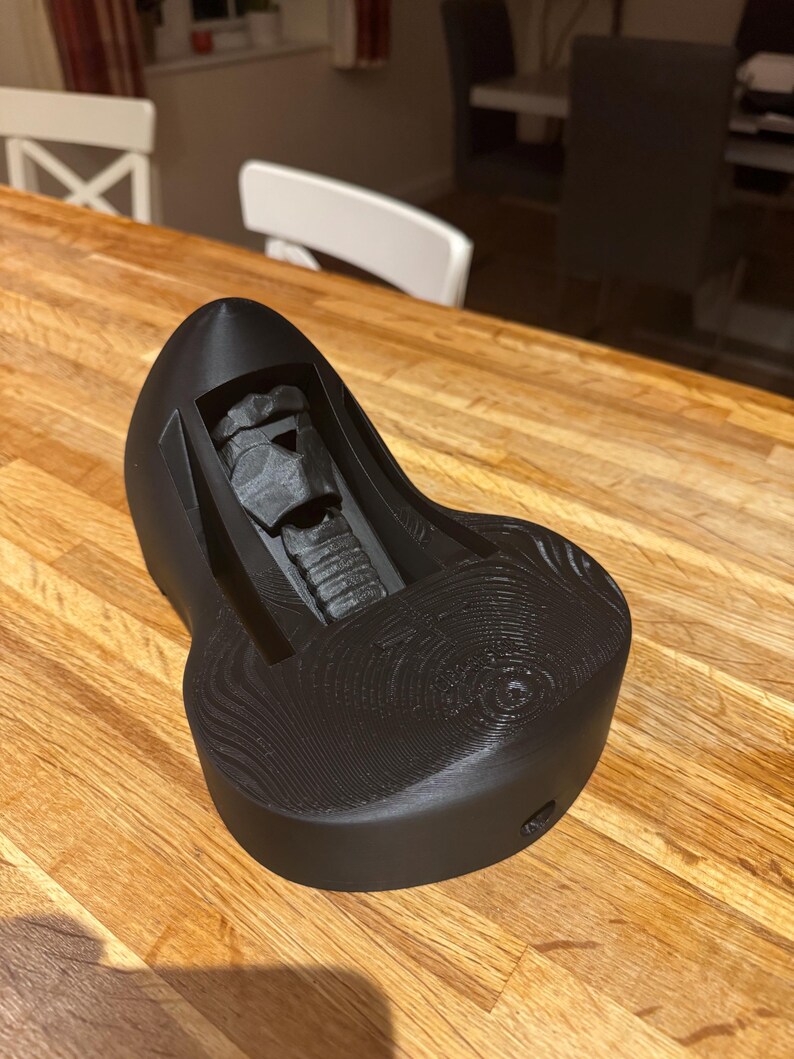 May include: A black, 3D-printed Darth Vader helmet-shaped phone holder. The holder has a textured surface and a slot to hold a phone. The design is detailed and the holder is sitting on a wooden table.