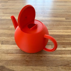 May include: A vibrant red teapot with a rounded body, a curved handle, and a spout. The lid is open, revealing the interior. The teapot is made of a solid material and sits on a wooden surface.