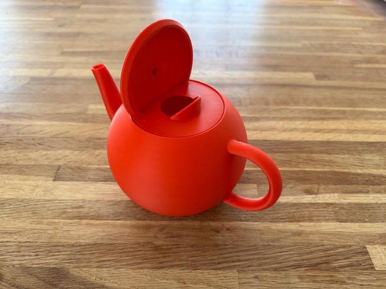 May include: A vibrant red teapot with a rounded body, a curved handle, and a spout. The lid is open, revealing the interior. The teapot is made of a solid material and sits on a wooden surface.