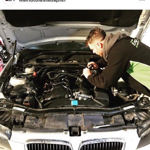 May include: A mechanic works on the engine of a white BMW car with the hood open. The engine is visible and the mechanic is wearing a black jacket and green shirt.