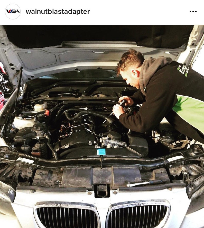 May include: A mechanic works on the engine of a white BMW car with the hood open. The engine is visible and the mechanic is wearing a black jacket and green shirt.