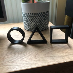 May include: Three black geometric shapes, a circle, a triangle, and a square, are arranged on a wooden surface. The shapes are made of a smooth, matte material.