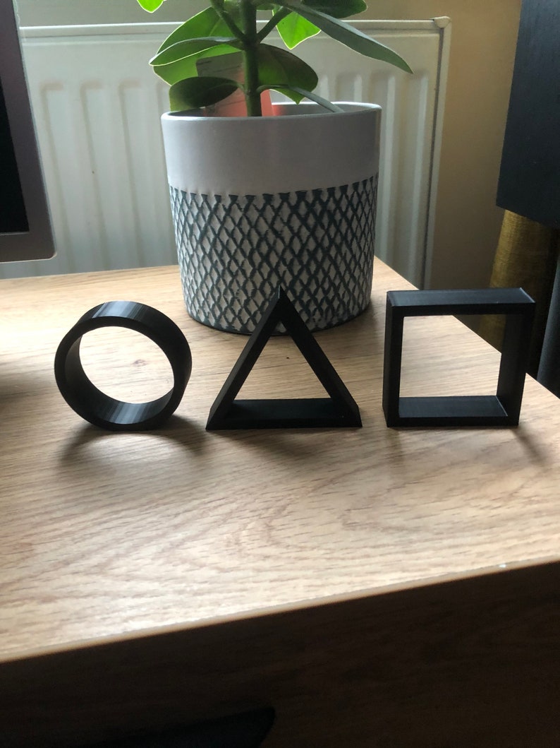 May include: Three black geometric shapes, a circle, a triangle, and a square, are arranged on a wooden surface. The shapes are made of a smooth, matte material.