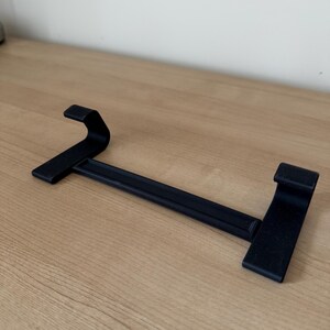 May include: A black metal shelf bracket with two upward-curving ends and a horizontal bar. The bracket is designed to hold a shelf and is made of a sturdy material. The bracket is shown on a light brown wooden surface.