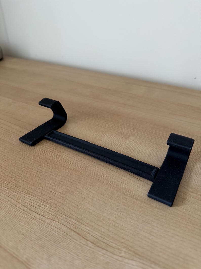 May include: A black metal shelf bracket with two upward-curving ends and a horizontal bar. The bracket is designed to hold a shelf and is made of a sturdy material. The bracket is shown on a light brown wooden surface.