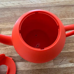 May include: A bright red watering can with a rounded body, a spout, and a handle. The can is made of plastic and appears to be empty. The lid is detached and lying next to the can. The watering can is sitting on a wooden surface.
