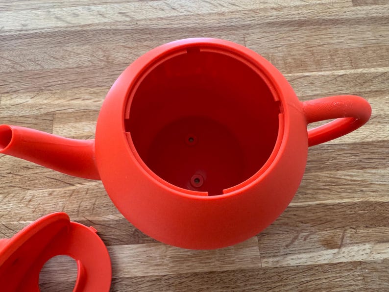 May include: A bright red watering can with a rounded body, a spout, and a handle. The can is made of plastic and appears to be empty. The lid is detached and lying next to the can. The watering can is sitting on a wooden surface.