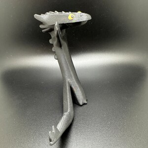 May include: Black 3D printed dragon figurine with yellow eyes. The dragon is posed in a standing position with its head tilted back and its wings spread out.