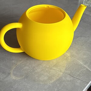 May include: A bright yellow teapot with a rounded body, a curved handle, and a spout. The teapot is made of a solid material and sits on a gray surface. The design is modern and minimalist.