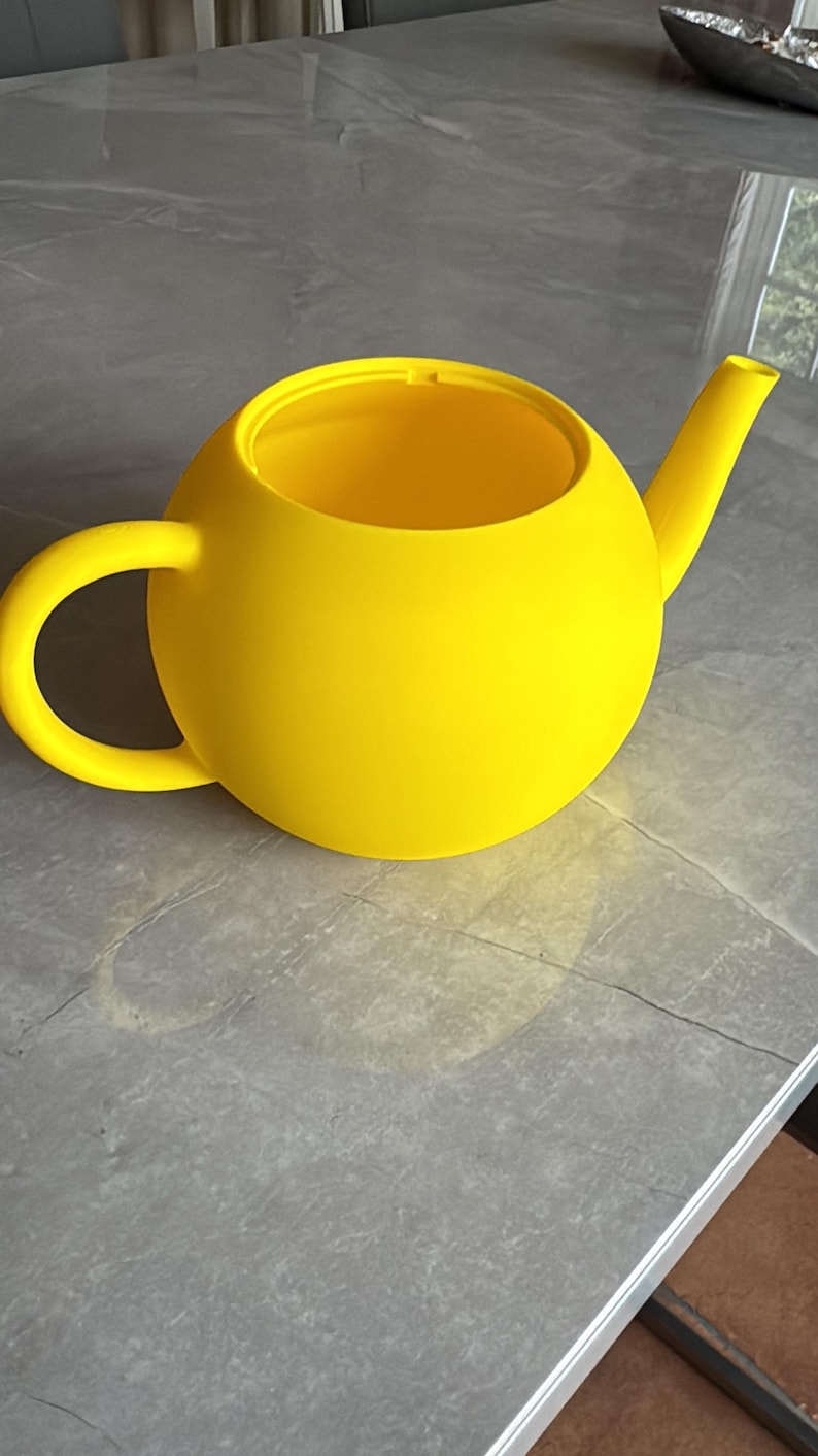 May include: A bright yellow teapot with a rounded body, a curved handle, and a spout. The teapot is made of a solid material and sits on a gray surface. The design is modern and minimalist.