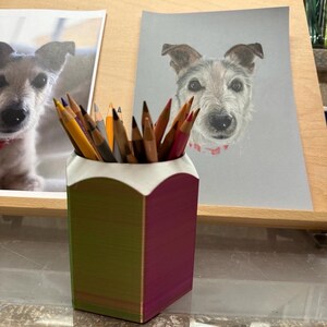 May include: A 3D printed pencil holder with a green and purple iridescent finish. The holder is filled with coloured pencils and is sitting on a wooden surface.