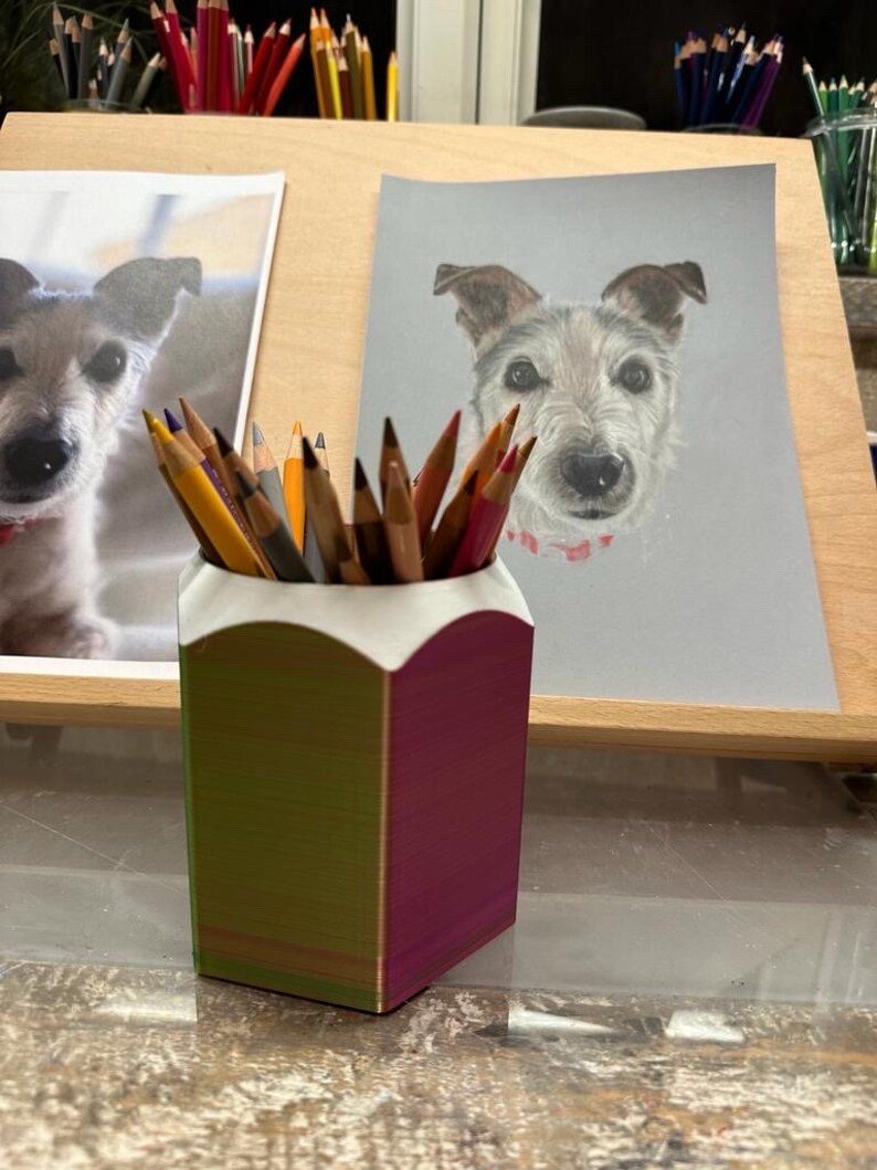 May include: A 3D printed pencil holder with a green and purple iridescent finish. The holder is filled with coloured pencils and is sitting on a wooden surface.