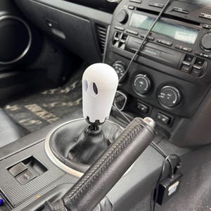 May include: A white ghost-shaped gear stick knob in a car. The knob has two eye holes and a round top. The car's interior is black, with a black leather-wrapped handbrake and a black centre console.