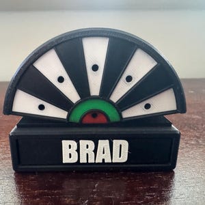 May include: Black and white dartboard with a green and red center. The dartboard is mounted on a black base with the name "BRAD" printed in white.