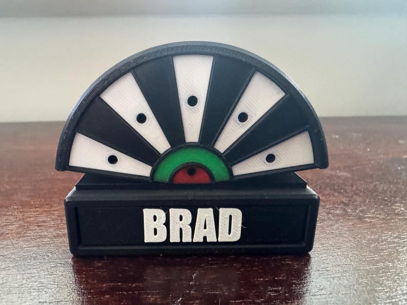 May include: Black and white dartboard with a green and red center. The dartboard is mounted on a black base with the name "BRAD" printed in white.