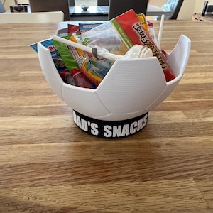 Op de afbeelding: Een witte, 3D-geprinte voetbalvormige kom gevuld met snoep en snacks. De kom staat op een zwarte basis met de tekst "DAD'S SNACKS" in het wit. De kom staat op een houten tafel.