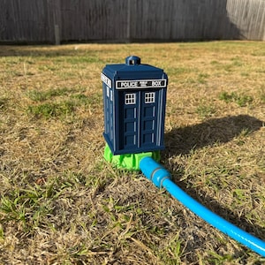 May include: A blue Police Box lawn sprinkler with white window details and the text "POLICE BOX". The sprinkler is connected to a blue hose and sits on a green base, placed on dry grass.