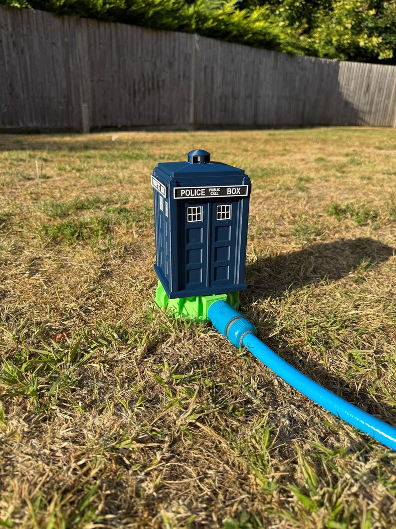 May include: A blue Police Box lawn sprinkler with white window details and the text "POLICE BOX". The sprinkler is connected to a blue hose and sits on a green base, placed on dry grass.