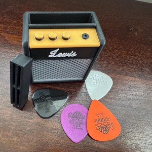 May include: A black and yellow miniature guitar amplifier with the word "Lewis" printed on the front. The amplifier has a slot for a phone or other device. Four guitar picks are in front of the amplifier, including one with the text "Jim Dunlop 111" and another with the text "Dunlop 460 USA".