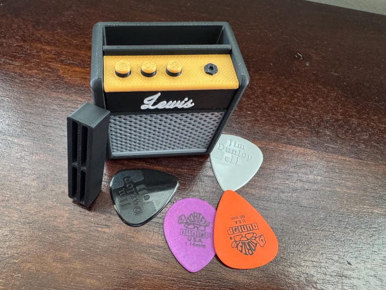 May include: A black and yellow miniature guitar amplifier with the word "Lewis" printed on the front. The amplifier has a slot for a phone or other device. Four guitar picks are in front of the amplifier, including one with the text "Jim Dunlop 111" and another with the text "Dunlop 460 USA".
