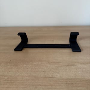 May include: A black, minimalist stand with a horizontal bar and two curved supports. The stand is designed to hold an item, with the supports providing stability. The object is made of a solid material and sits on a wooden surface.