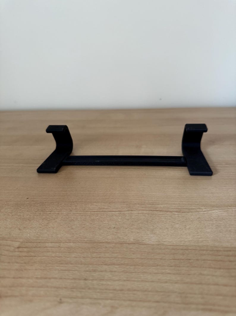 May include: A black, minimalist stand with a horizontal bar and two curved supports. The stand is designed to hold an item, with the supports providing stability. The object is made of a solid material and sits on a wooden surface.