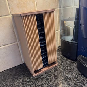 May include: A light brown, rectangular tea bag organizer with vertical slats on the front and sides. The organizer is open in the front, revealing several tea bags. A blue coffee maker and water dispenser are in the background.