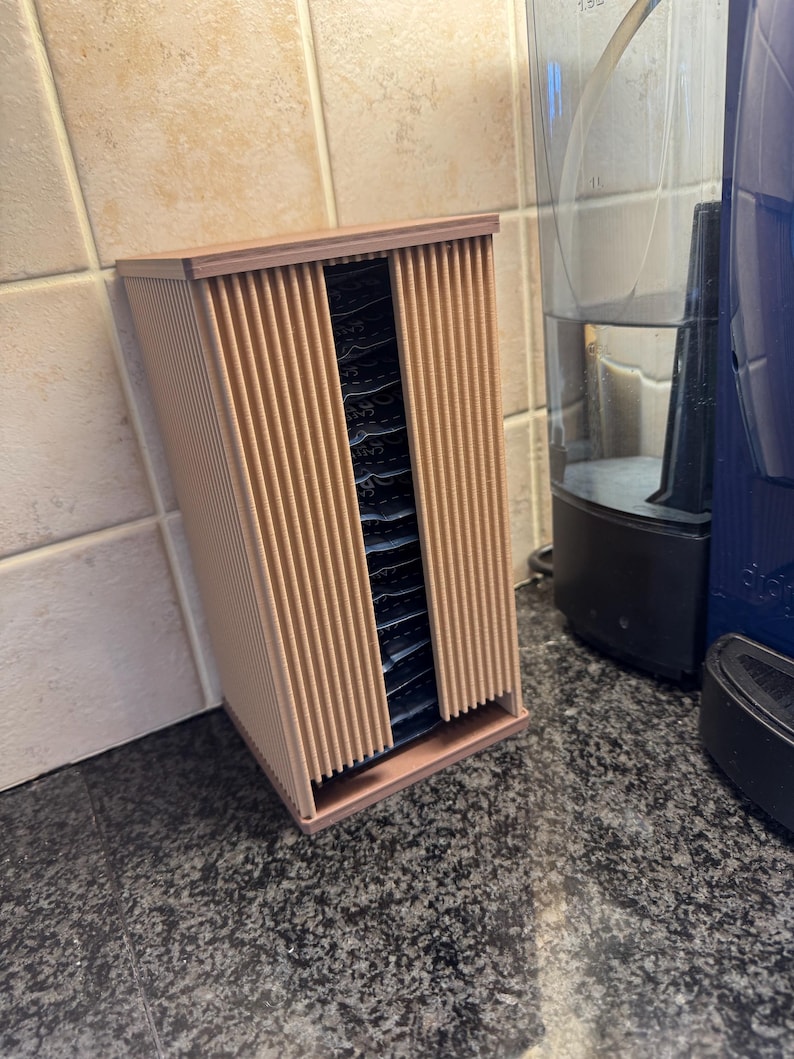 May include: A light brown, rectangular tea bag organizer with vertical slats on the front and sides. The organizer is open in the front, revealing several tea bags. A blue coffee maker and water dispenser are in the background.